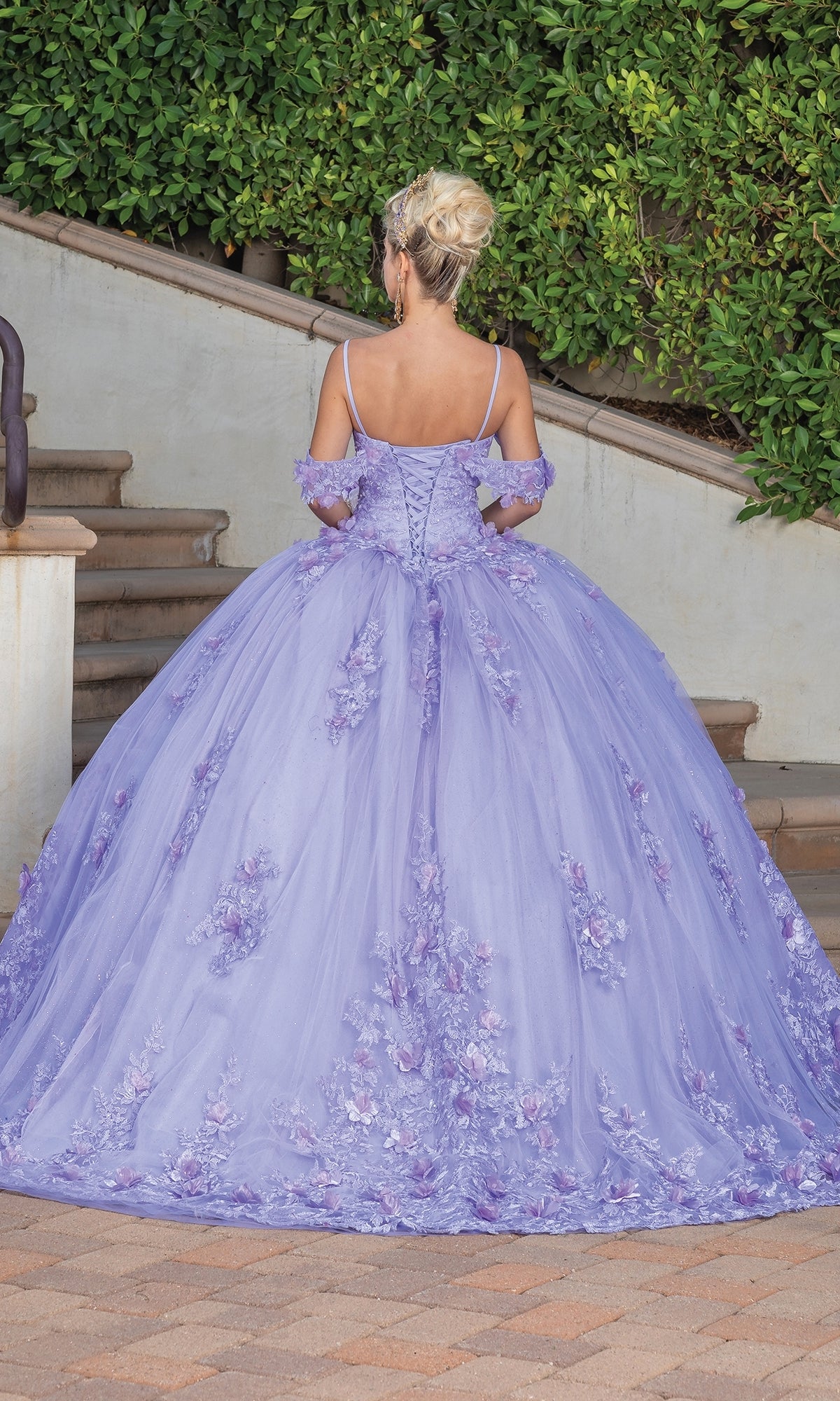 A woman with blonde hair in an updo wears the Quinceanera Dress 1747 By Dancing Queen, a lilac ball gown with floral details, while standing on an outdoor staircase surrounded by greenery.